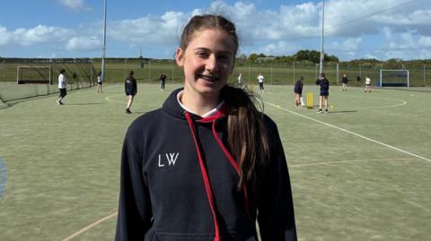 Lily, a pupil at Cockermouth School, stands in front of the astro turf cricket pitch wearing a sports hoodie.