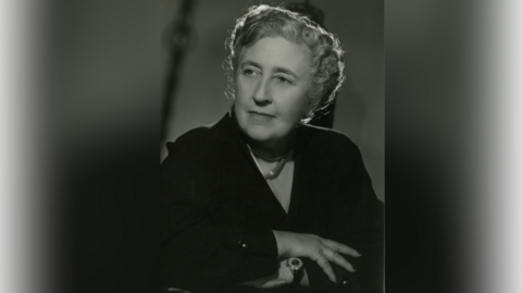 An archive sepia image of Agatha Christie, around 1950. She is sitting down with her arms folded and looking away from the camera.