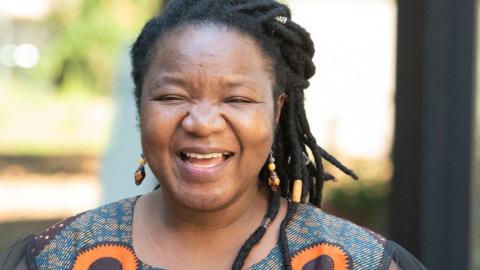 Angeline Murimirwa, smiling broadly towards the camera. She has pulled back dark hair and is wearing a blue, orange and brown top and yellow brown and blue earrings