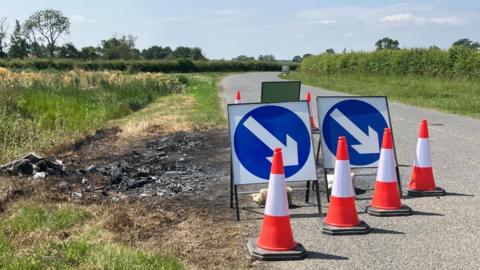 A series of cones and road signs send drivers around a scorched patch of earth at the side of a country road