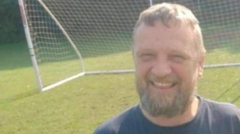 Robin Reed smiling for the camera. He has a grey beard and is wearing a navy blue t-shirt that has the Labretta brand. Behind him is a football goalpost.