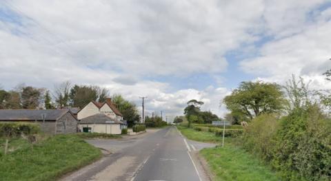 Road junction looking with a small lane running left to right there are road signs on the left side.  To the left is a barn and other buildings.