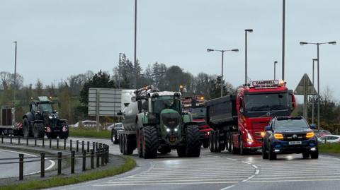 A convoy of trucks and tractors driving down a road on an overcast day.