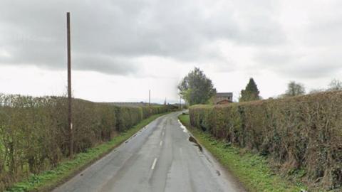 A narrow road with hedges on both sides and a pole on the left hand side under a grey sky
