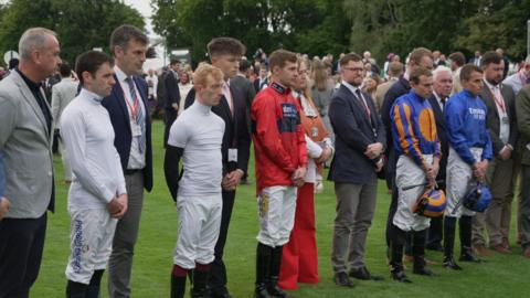 Staff and jockeys stand in a line in silence