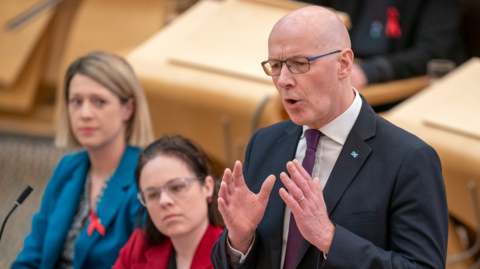 John Swinney answering questions in the Holyrood chamber as he stands next to Kate Forbes in red suit and Jenny Gilruth in blue suit