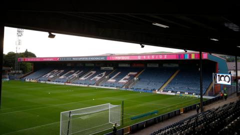 A view from behind the goal at Rochdale's Crown Oil Arena, with a view of the stand along the touchline with 'ROCHDALE' painted across the seats. 
