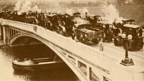 A black and white concrete bridge with one person standing on it next to many steam-powered vehicles all parked on it. 