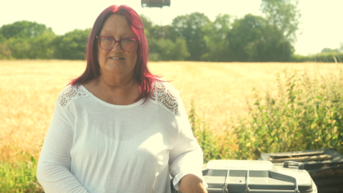 A woman with red hair wearing a white T-shirt standing in front of a field. The tops of bins are to the right of the frame.