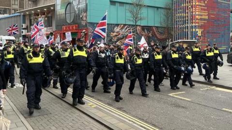 A small group of people march with Union flags and england flags while being escorted by a large group of police