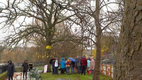 A number of people gathered around an oak tree. It is raining, with some people carrying umbrellas. 