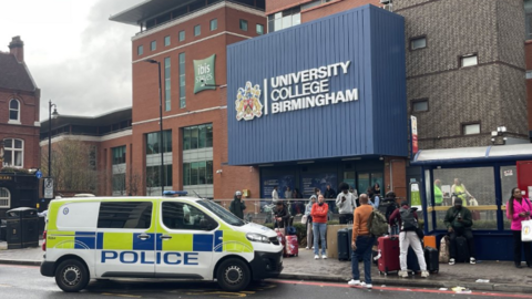 A police van parked outside a college building with a big blue sign saying 'University College Birmingham'. There are around a dozen people outside the building which also has a bus shelter in front of it.