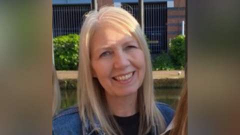 Shelley Hallam has long blonde hair and blue eyes. She is wearing a blue denim jacket.