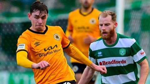 Matty Carson in his orange Boston United kit passes the ball under pressure from a Yeovil Town player wearing a green and white hooped shirt. There is another Yeovil player in the background and another Boston one in the distance.