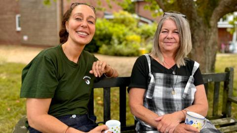 Two women sitting on a parking bench, both holding mugs of tea, with both looking to the camera. One has dark hair and is wearing a dark green T-shirt, while the other has blonde hair and is wearing black and white check dungarees over a black T-shirt.