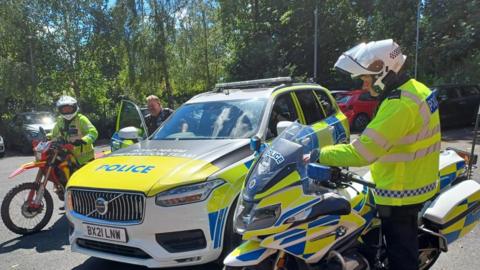 A blue, white and yellow police car is parked and there are three police officers next to it. On the right there is a policeman in a yellow fluorescent jacket and white helmet. He is standing next to a yellow, white and blue police motorbike. On the left are two police officers. One is wearing all black police uniform and is getting into the driver's side door which is open. Next to him is another police officer who is holding a red and yellow off-road bike. The officer is wearing yellow fluorescent gear and white helmet