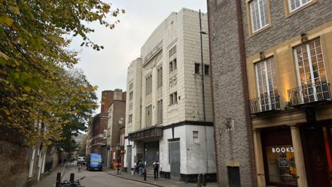 Hobson Street, with the former cinema on the right-hand side. You can see the building's art deco facade but the brickwork is discoloured and the front doors and windows are boarded up. Next to it is a bookshop.