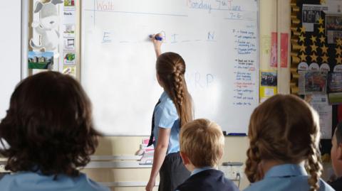 A young girl is writing on a whiteboard in front of a class, all of whom have their backs to the camera.