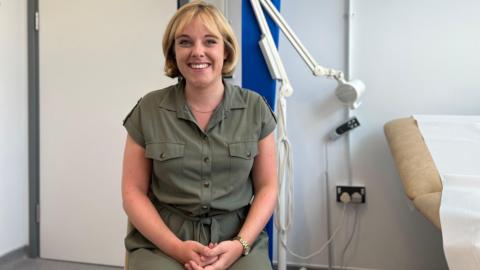 Georgia smiles at the camera as she sits on chair next to an empty patient bed