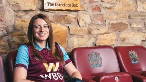 Justine Bedford with long dark hair wearing a Burnley FC shirt sitting on leather Claret chairs with the club logo on in her pub with a wooden sign above her which reads: The Directors Box. She is smiling.