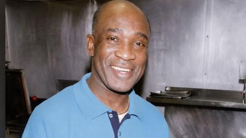 A close-up of Alfanso Vivian Salmon smiling. He has no hair and is wearing a blue polo shirt.