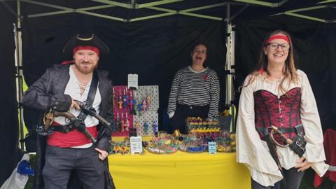 A man and a woman dressed as pirates smile in front of a stall at the rum butter festival. A female retailer a black and white striped top is smiling behind a table. 
