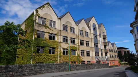 A yellow building wikth six triangular pointed roofs. The lower left third of the building is covered in green ivy. 