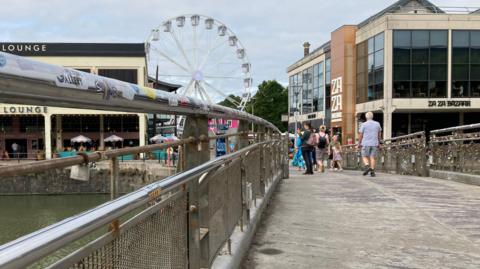 Pero's Bridge in Bristol. There are railways either side of the pathway. There are some padlocks hanging on it. In the distance, people can be seen walking around and there are restaurants and a white ferris wheel.