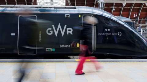 A dark green Great Western Railway train is parked by a platform. People are walking past.