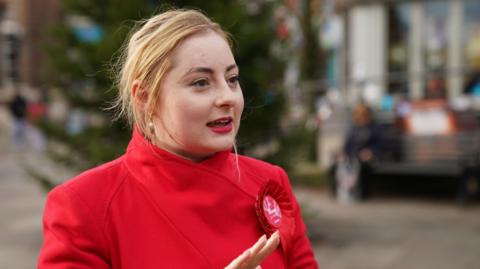 A blonde woman in a red coat and red rosette speaks side on to the camera outside in a town centre.