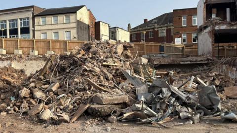 A pile of building rubble with concrete and metal, awaiting recycling. The pile sits in front of a wooden fence which is in front of several buildings.