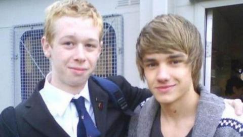 John Carpenter has blonde short hair and is wearing a black blazer, blue tie and white shirt. He is standing next to Liam Payne who has short brown hair and is wearing a grey cardigan.