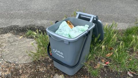 A "Suffolk recycles" food caddy is on the kerb with pavement and grass visible either side. The caddy is grey with a black handle and is full of food waste. 