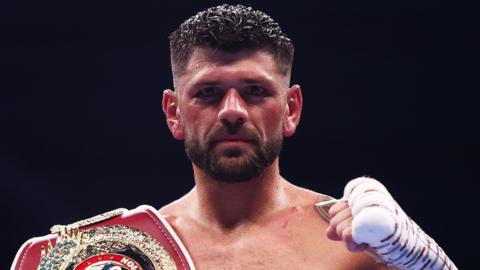 Joe Cordina poses with bandaged left fist raised and boxing belt over right shoulder