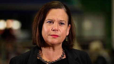 Mary Lou McDonald looking to camera wearing a black suit and necklace