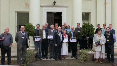 A group photograph showing the award winners and council dignitaries holding their certificates, standing outside a white building.