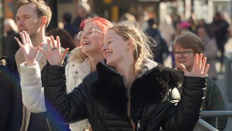 Two young women in a crowd singing and waving their hands to the musical performance. 