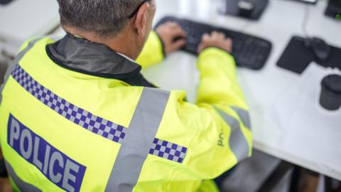 A male police officer sat at a computer, typing. He wears a high-vis jacket with "police" written on it. He has short, dark or grey hair and has the end of a spectacle limb visible over one ear.