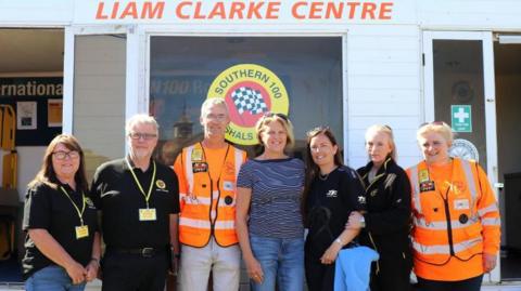 People who knew Liam Clarke line up outside the centre. Two marshals wear orange uniforms. The sign reads Liam Clarke Centre in orange writing on a white exterior wall.