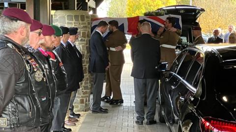 Image shows a coffin being carried into a funeral. The coffin has a union jack flag draped over it and is being carried by men in military uniform. More men in military berets are stood paying respects as the coffin is being carried into the venue from the lined up black cars.