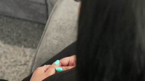 A woman is seen from behind and above. She has long dark hair, is sitting on a grey sofa and holds her hands together on her lap. Her nails are painted a bright turquoise.