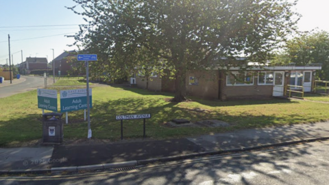 A street sign in Beverley reading "Coltman Avenue". The sign is at the side of a road underneath a large tree. An Adult Learning Centre is in the background.