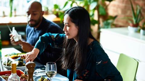 A woman and man sit side-by-side with plates of food in front of them. The woman is serving herself from a dish in front of her.