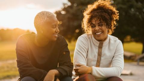 Two women sitting in a park setting. Chatting and smiling. Sunset in the back ground. One is wearing black the other is is wearing white. 