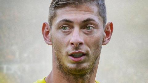 Emiliano Sala has short brown hair and is wearing a yellow Nantes Football Club top. Crowds of spectators at a football ground are blurred out behind him.