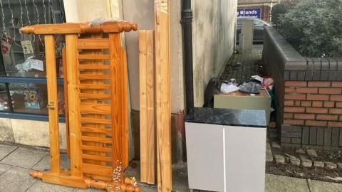 A wooden bed frame, drawers and bags of rubbish can be seen in an alleyway.