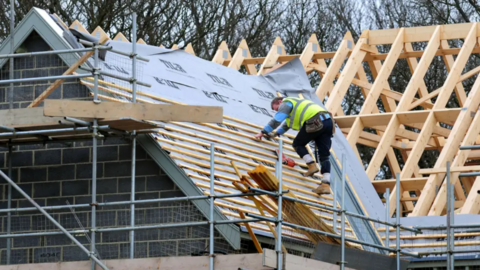 A wide shot of a man climbing on a roof which is being supported by scaffolding. He is wearing a high vis jacket.