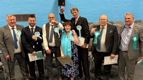 Seven Reform UK politicians are seen smiling and celebrating. They are wearing light blue rosettes and are stood against a blue wall.