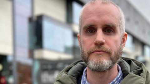 Aaron Wyllie stands in front of The Forum building in Southend-on-Sea. He is wearing a green jacket over a black jumper and blue shirt. He has a beard and short cut hair.