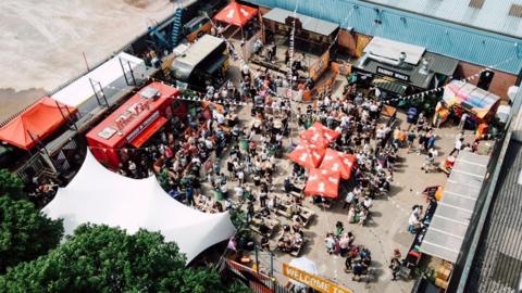 An aerial shot of a many food stalls with dozens of people milling about with some sat at wooden tables in the daytime.